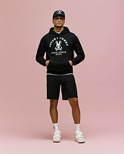A man stands against a pink background wearing a black Psycho Bunny MENS ARCHIVE HOODIE (MT0500009), black cap, black shorts, white socks, and white sneakers, with his hands in the hoodie pockets.