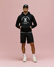 A man stands against a pink background wearing a black Psycho Bunny MENS ARCHIVE HOODIE (MT0500009), black cap, black shorts, white socks, and white sneakers, with his hands in the hoodie pockets.