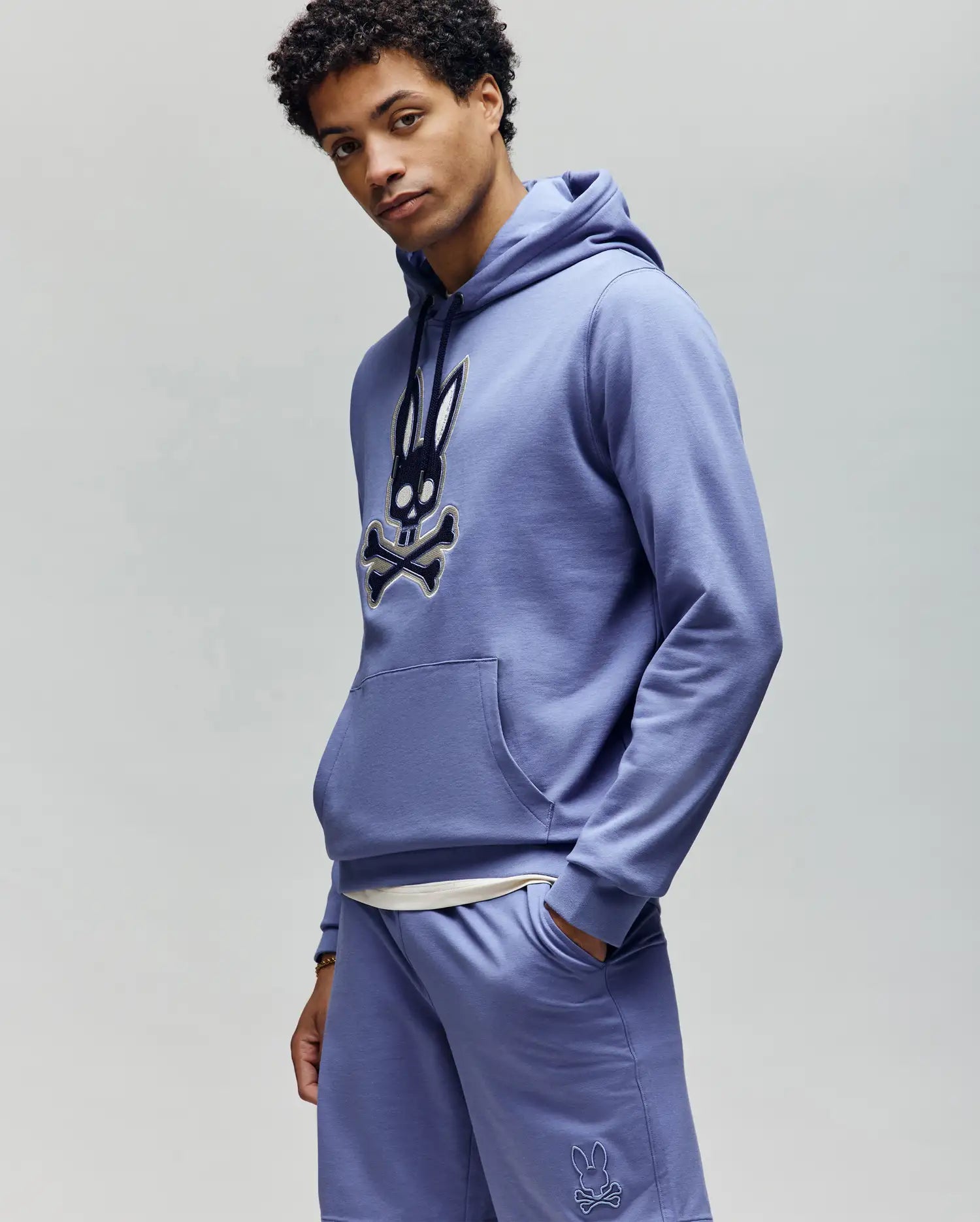 A man faces forward in a lavender Psycho Bunny MENS RYDER EMBROIDERED BUNNY PATCH HOODIE (MT0500029) and matching shorts, featuring a bold embroidered skull and crossbones bunny on the front. The background is plain light gray.