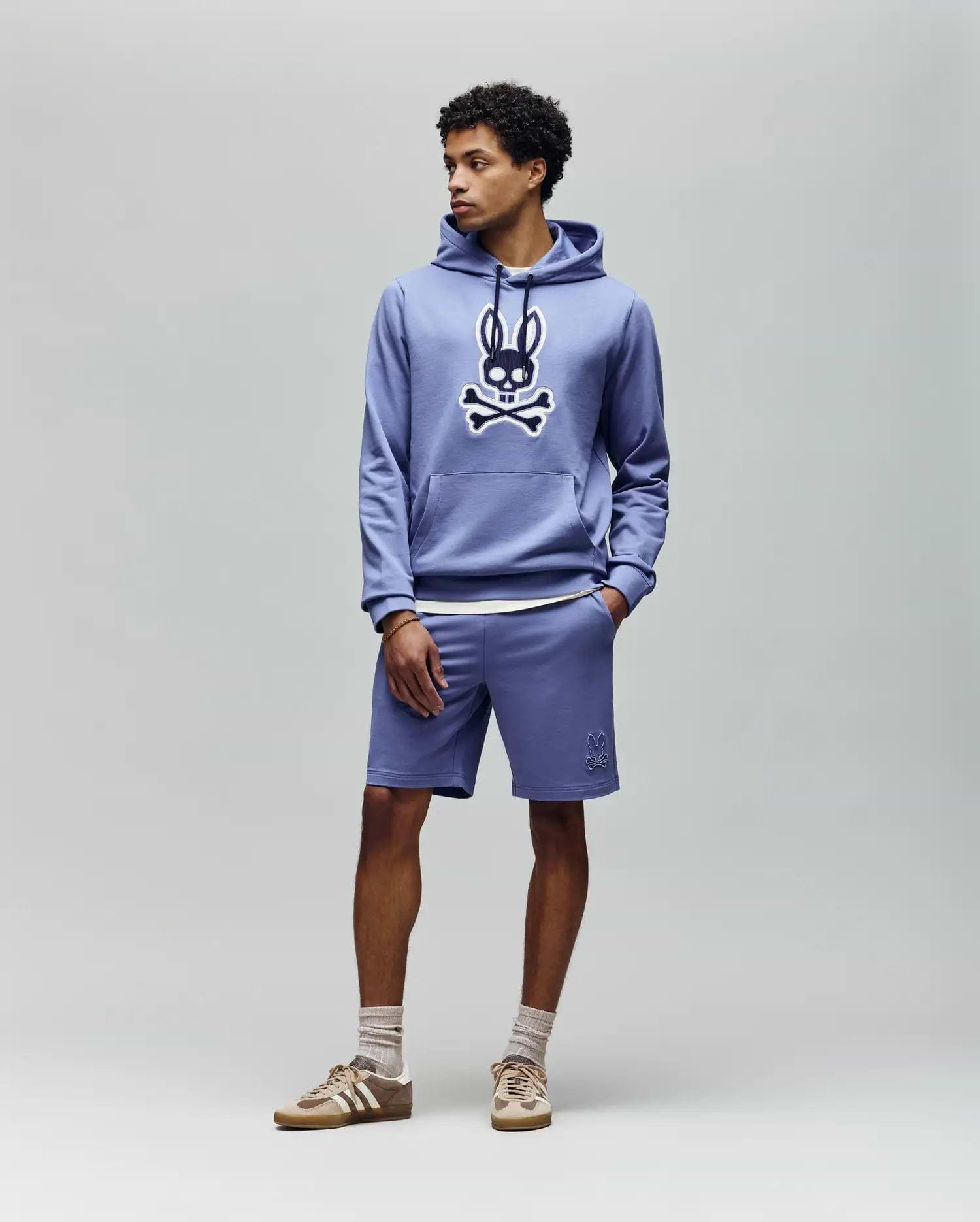 A man wears the Psycho Bunny MENS RYDER EMBROIDERED BUNNY PATCH HOODIE (MT0500029) in light blue, paired with matching shorts, beige socks, and beige sneakers, standing against a plain light gray background.