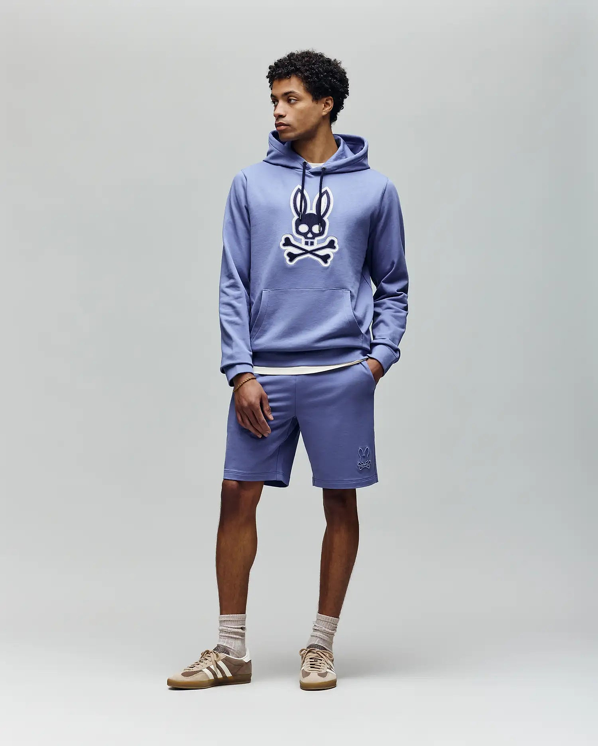 A man wears the Psycho Bunny MENS RYDER EMBROIDERED BUNNY PATCH HOODIE (MT0500029) in light blue, paired with matching shorts, beige socks, and beige sneakers, standing against a plain light gray background.