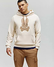 A man wears the Psycho Bunny MENS RYDER EMBROIDERED BUNNY PATCH HOODIE (MT0500029) in light beige, featuring an embroidered Bunny patch and crossbones on the front, styled with tan cargo pants against a plain light gray background.
