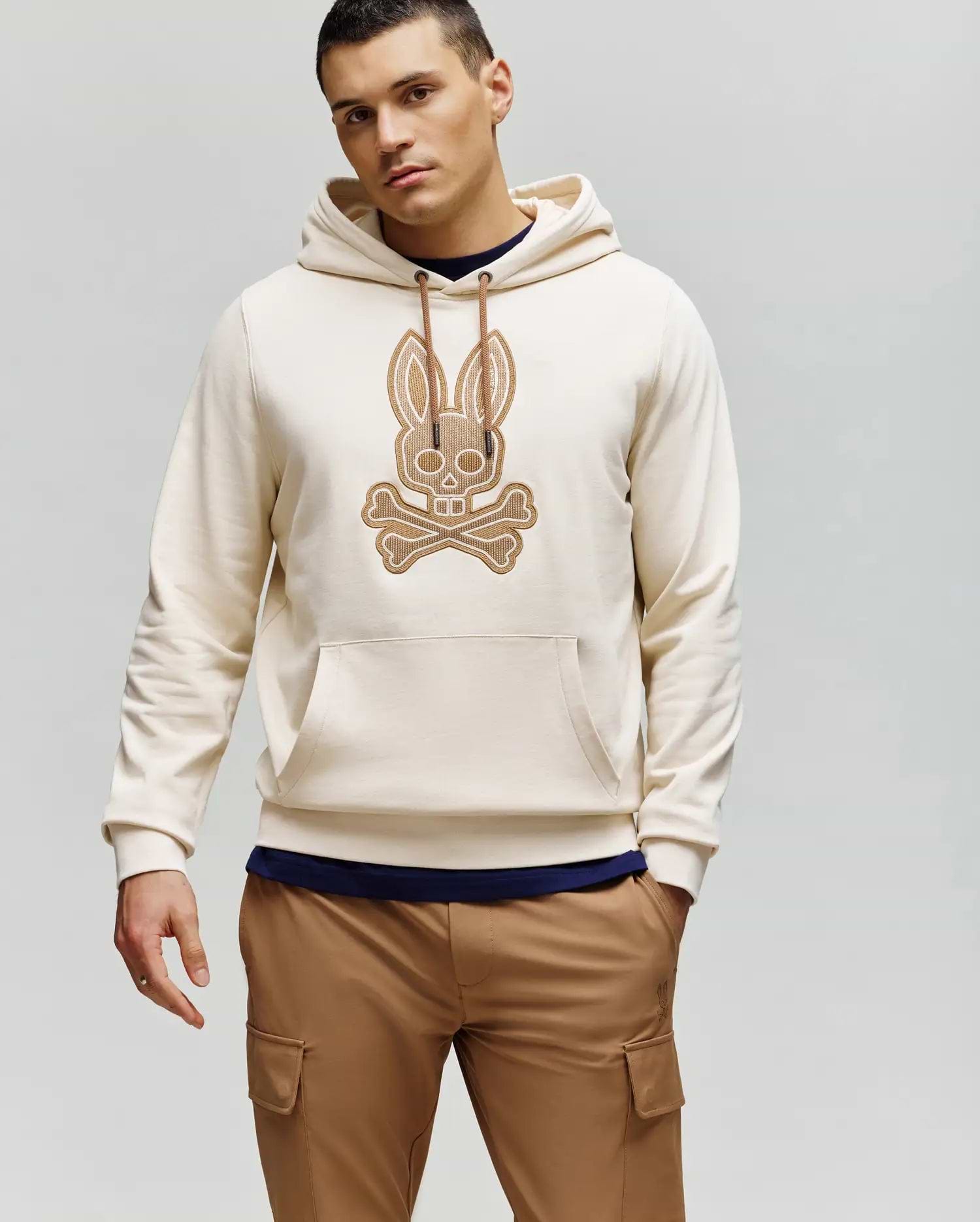 A man wears the Psycho Bunny MENS RYDER EMBROIDERED BUNNY PATCH HOODIE (MT0500029) in light beige, featuring an embroidered Bunny patch and crossbones on the front, styled with tan cargo pants against a plain light gray background.