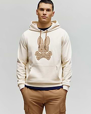 A man with short dark hair stands against a plain light background, wearing the Psycho Bunny MENS RYDER EMBROIDERED BUNNY PATCH HOODIE (MT0500029) in beige with a large crossbones graphic, paired with brown cargo loungewear pants.