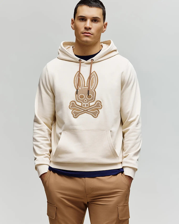 A man with short dark hair stands against a plain light background, wearing the Psycho Bunny MENS RYDER EMBROIDERED BUNNY PATCH HOODIE (MT0500029) in beige with a large crossbones graphic, paired with brown cargo loungewear pants.