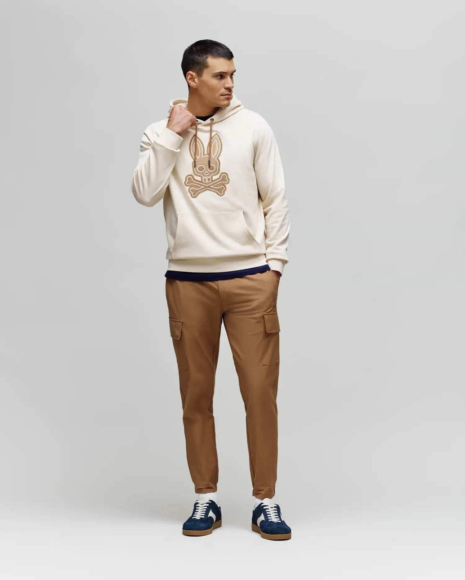 A man stands against a plain background wearing the Psycho Bunny MENS RYDER EMBROIDERED BUNNY PATCH HOODIE (MT0500029), paired with tan cargo pants and navy blue sneakers—ideal for a relaxed, laid-back look.