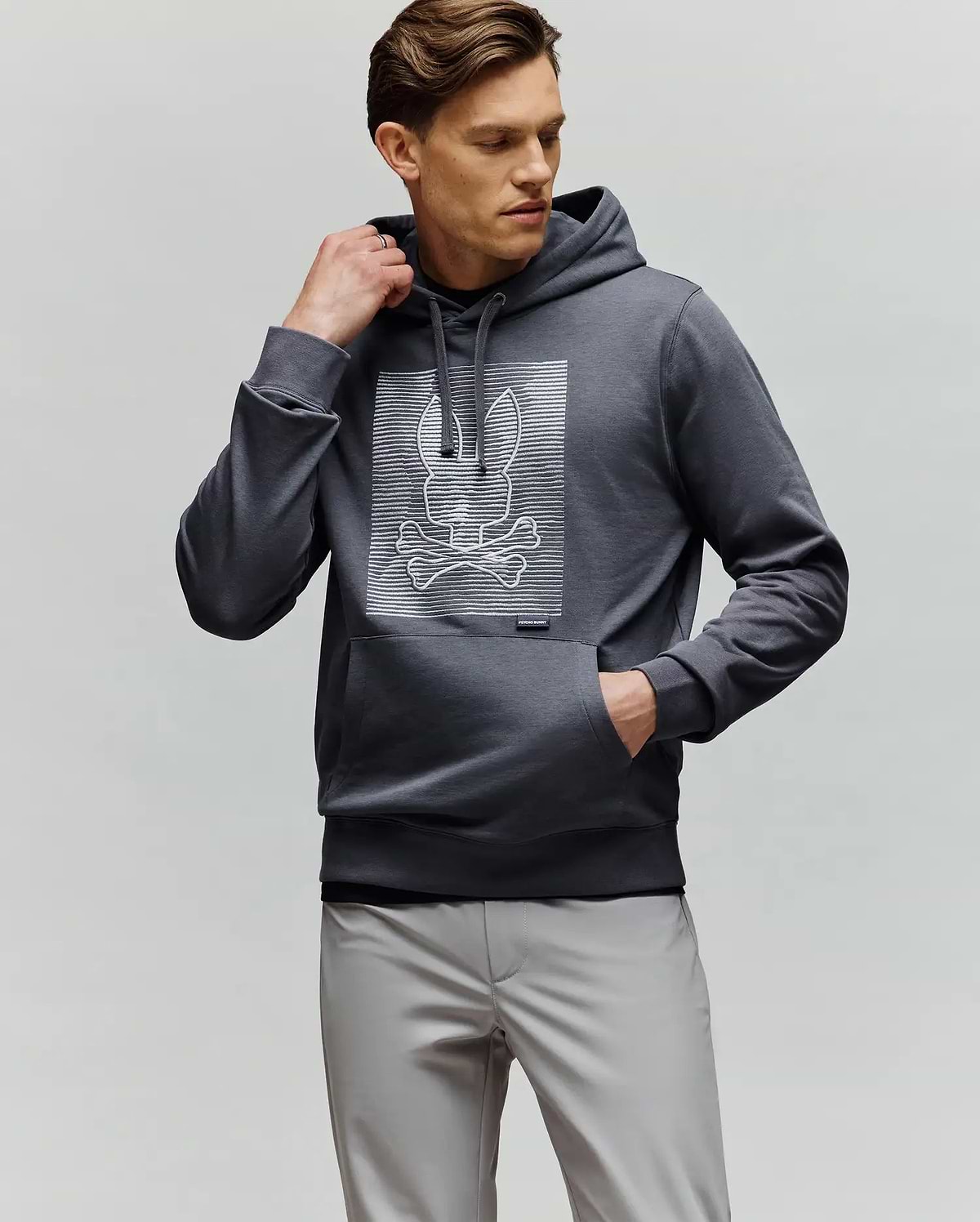 A man wears the Psycho Bunny MENS GRANT HOODIE - MT0500032, a dark gray premium cotton hoodie featuring a digital-inspired bunny and crossbones graphic. He styles it with light gray pants, one hand in his pocket and looking to the side.