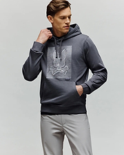 A man wears the Psycho Bunny MENS GRANT HOODIE - MT0500032, a dark gray premium cotton hoodie featuring a digital-inspired bunny and crossbones graphic. He styles it with light gray pants, one hand in his pocket and looking to the side.