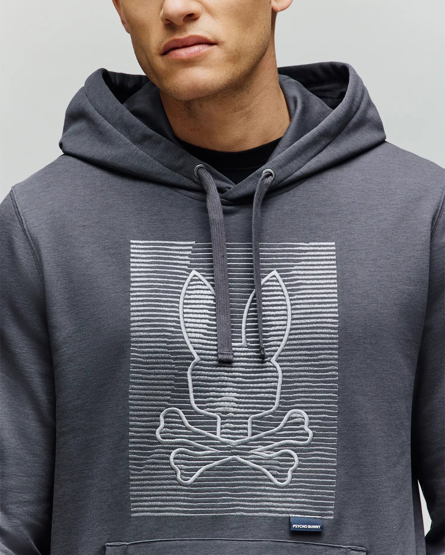 A person wears the Psycho Bunny MENS GRANT HOODIE (MT0500032), featuring a premium cotton build with an embroidered bunny head above crossed bones, inspired by the skull and crossbones, set against a backdrop of digital-style horizontal lines.