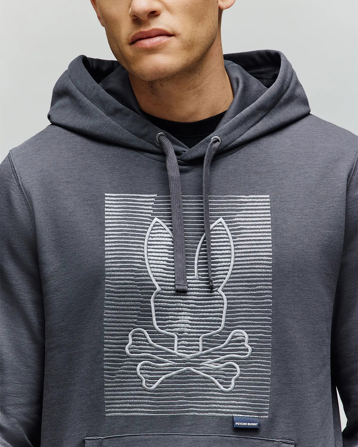 A person wears the Psycho Bunny MENS GRANT HOODIE (MT0500032), featuring a premium cotton build with an embroidered bunny head above crossed bones, inspired by the skull and crossbones, set against a backdrop of digital-style horizontal lines.