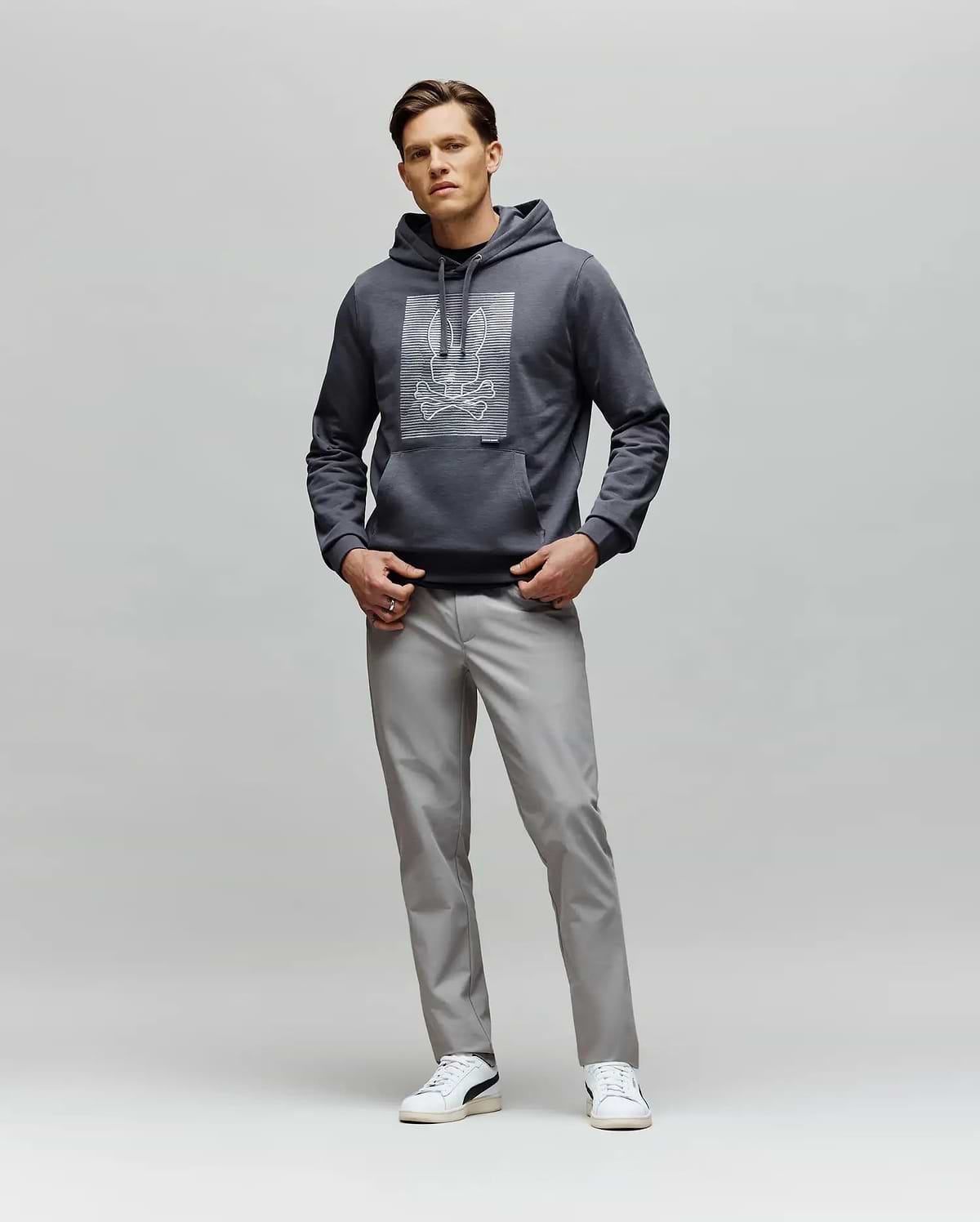 A man faces forward wearing the Psycho Bunny MENS GRANT HOODIE - MT0500032 with a digital-inspired graphic, paired with light gray pants and white sneakers, set against a plain light gray background.