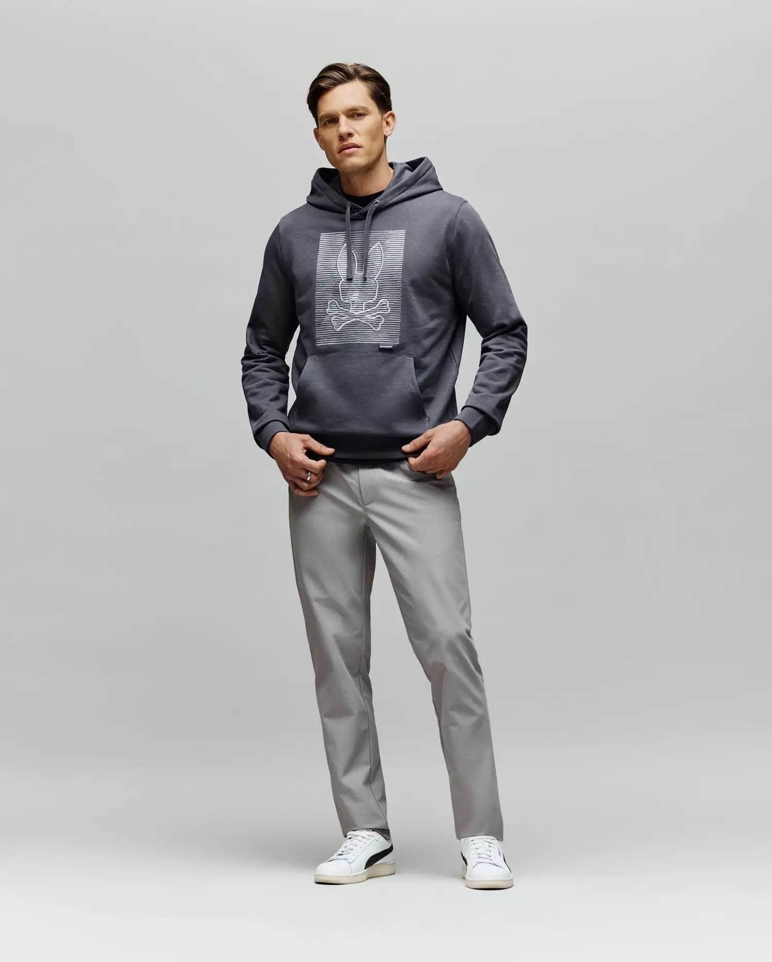 A man faces forward wearing the Psycho Bunny MENS GRANT HOODIE - MT0500032 with a digital-inspired graphic, paired with light gray pants and white sneakers, set against a plain light gray background.