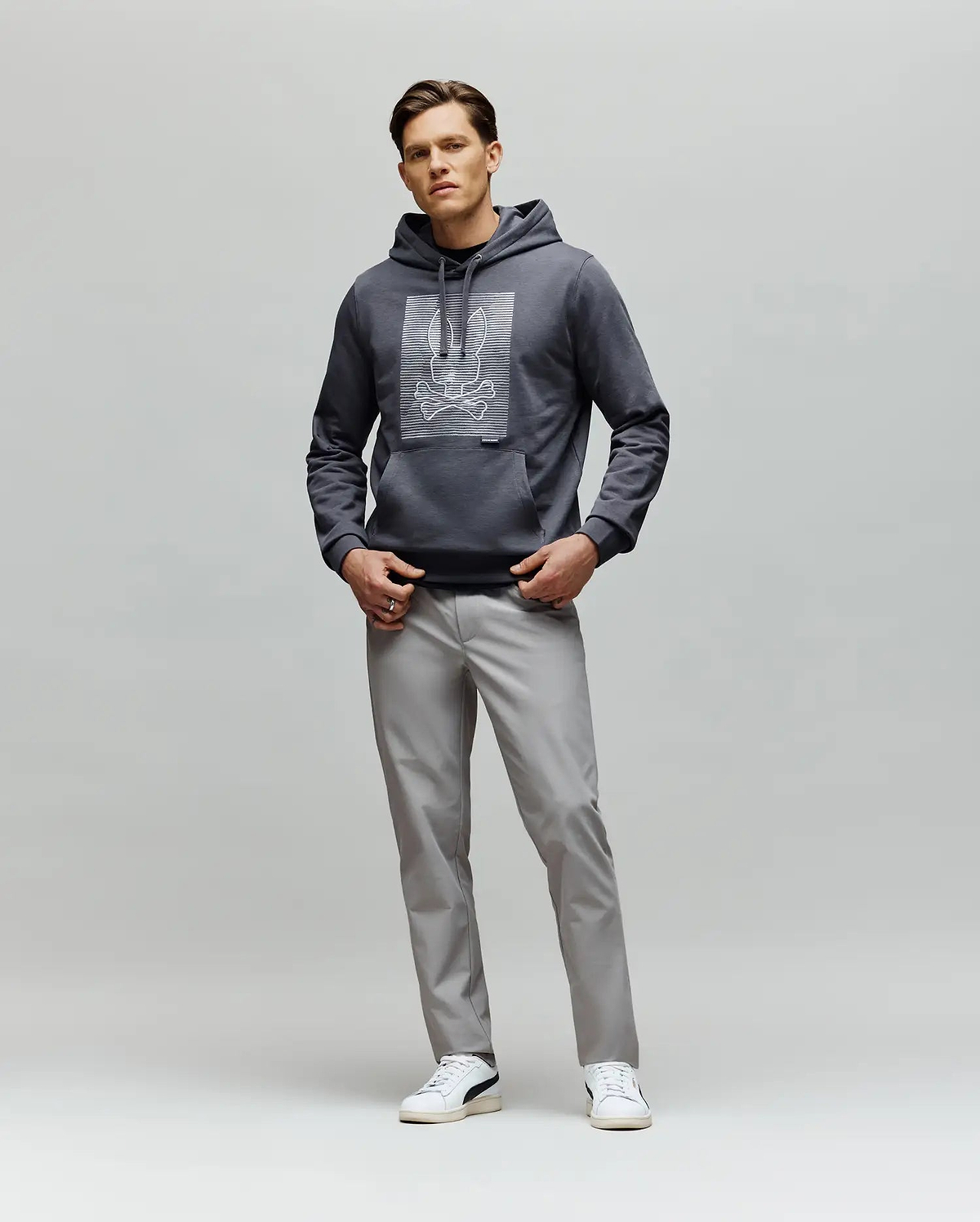 A man faces forward wearing the Psycho Bunny MENS GRANT HOODIE - MT0500032 with a digital-inspired graphic, paired with light gray pants and white sneakers, set against a plain light gray background.