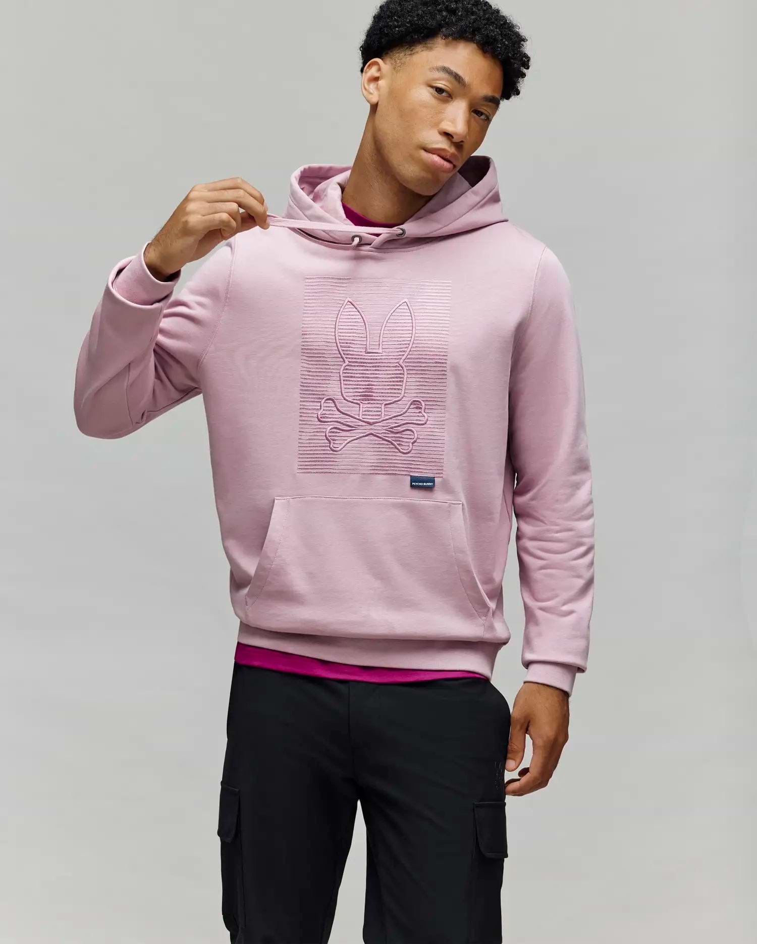 A man in a light pink Psycho Bunny MENS GRANT HOODIE (MT0500032), crafted from a premium cotton blend with a raised bunny and crossbones design, stands against a neutral background holding the hood, paired with black pants.