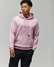 A man wears the Psycho Bunny MENS GRANT HOODIE - MT0500032 in light pink, crafted from a premium cotton blend with a raised skull and bunny ears design, standing against a plain gray background.