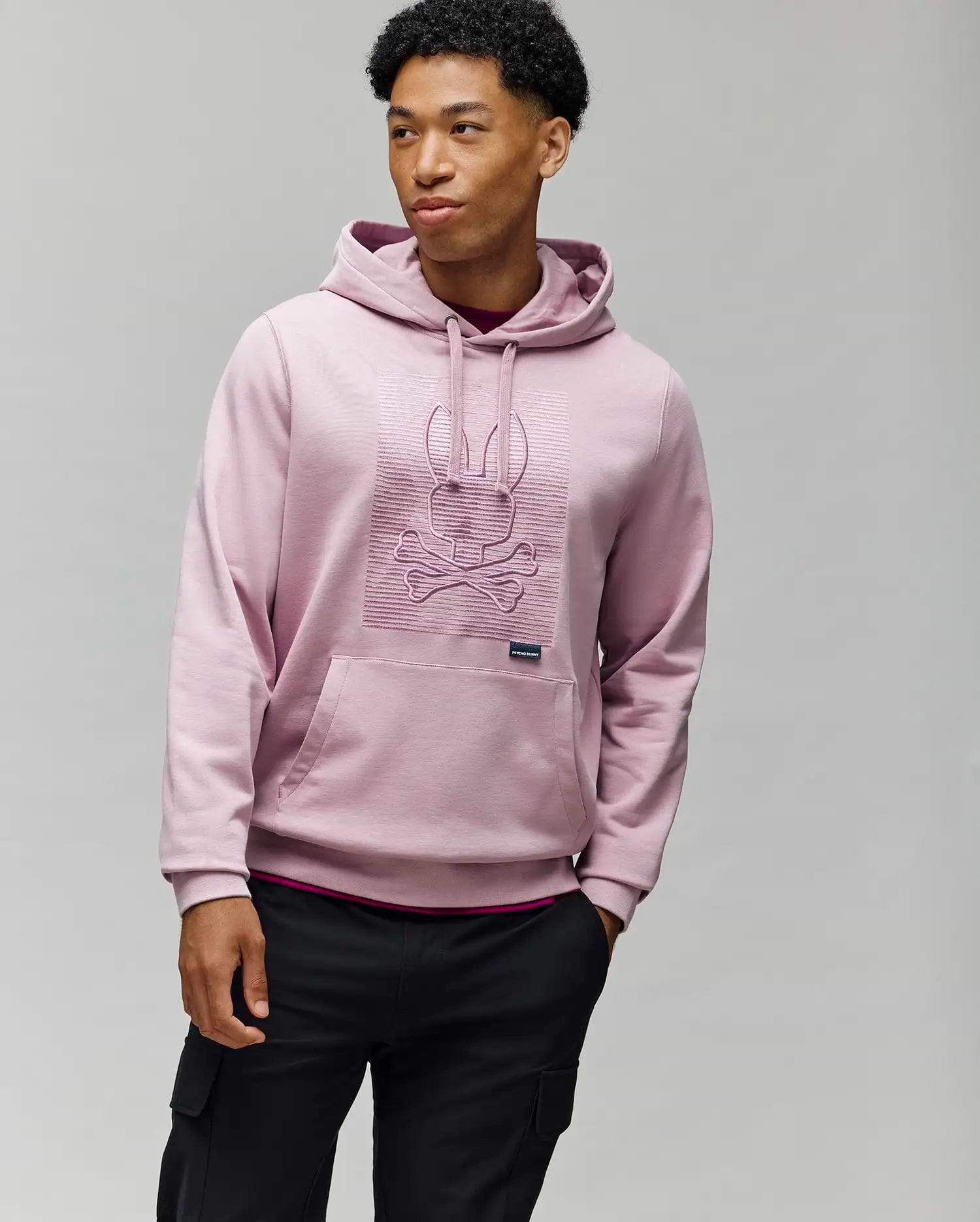 A man wears the Psycho Bunny MENS GRANT HOODIE - MT0500032 in light pink, crafted from a premium cotton blend with a raised skull and bunny ears design, standing against a plain gray background.