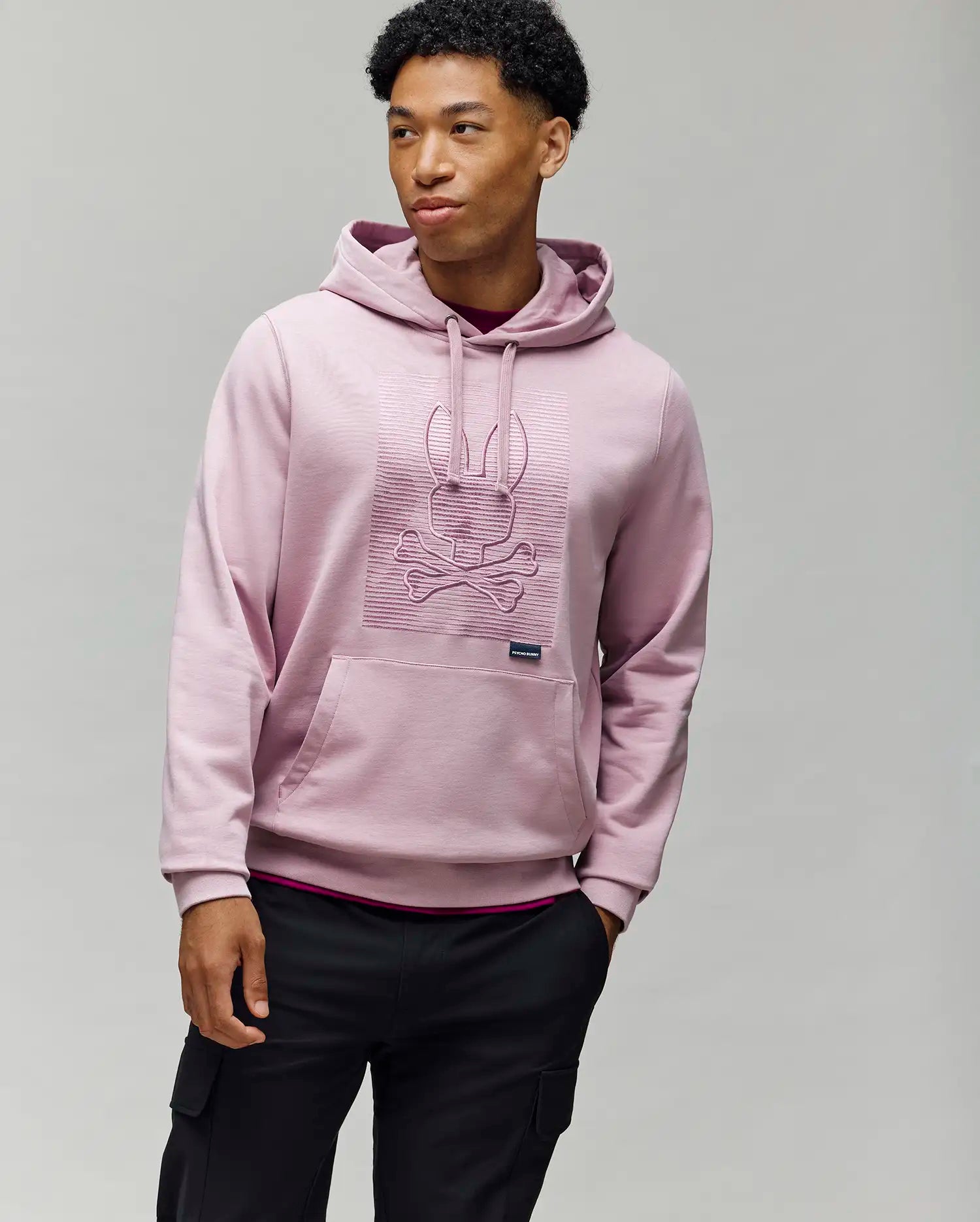 A man wears the Psycho Bunny MENS GRANT HOODIE - MT0500032 in light pink, crafted from a premium cotton blend with a raised skull and bunny ears design, standing against a plain gray background.