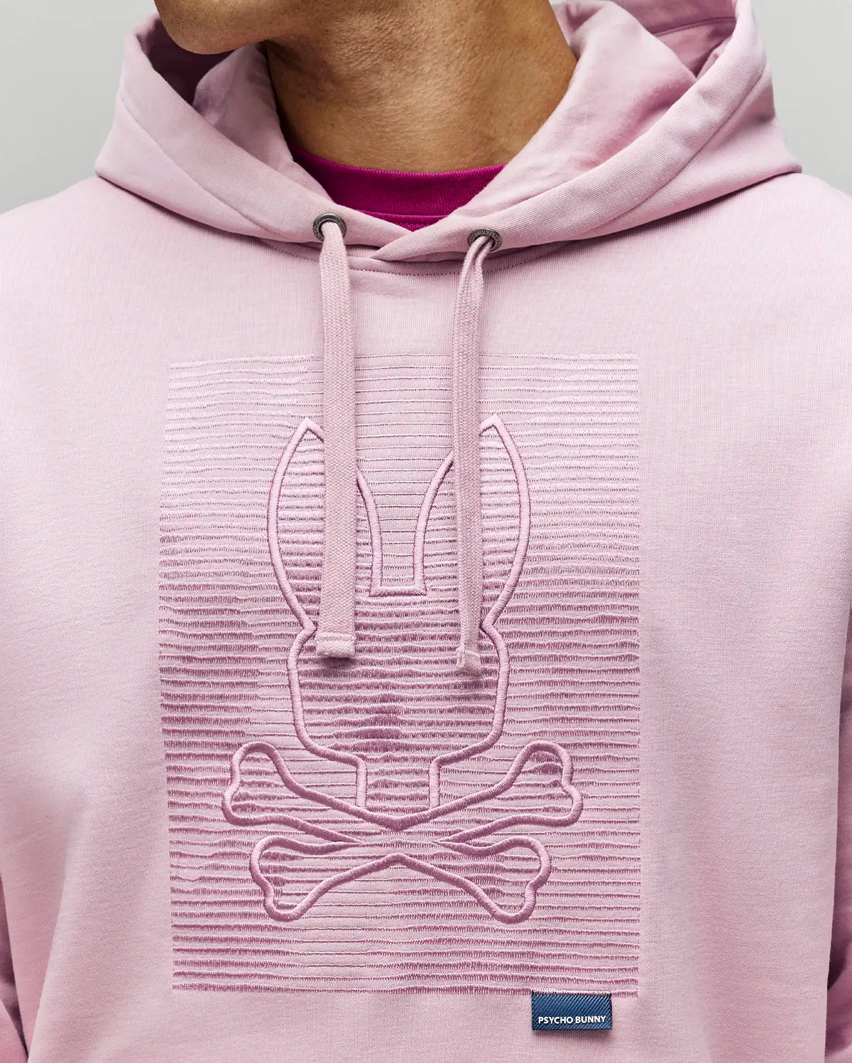 A person wears the Psycho Bunny MENS GRANT HOODIE - MT0500032 in light pink, made from a premium cotton blend, with an embossed bunny and crossbones on the chest and a "Psycho Bunny" label near the hem; only their lower face and neck show.