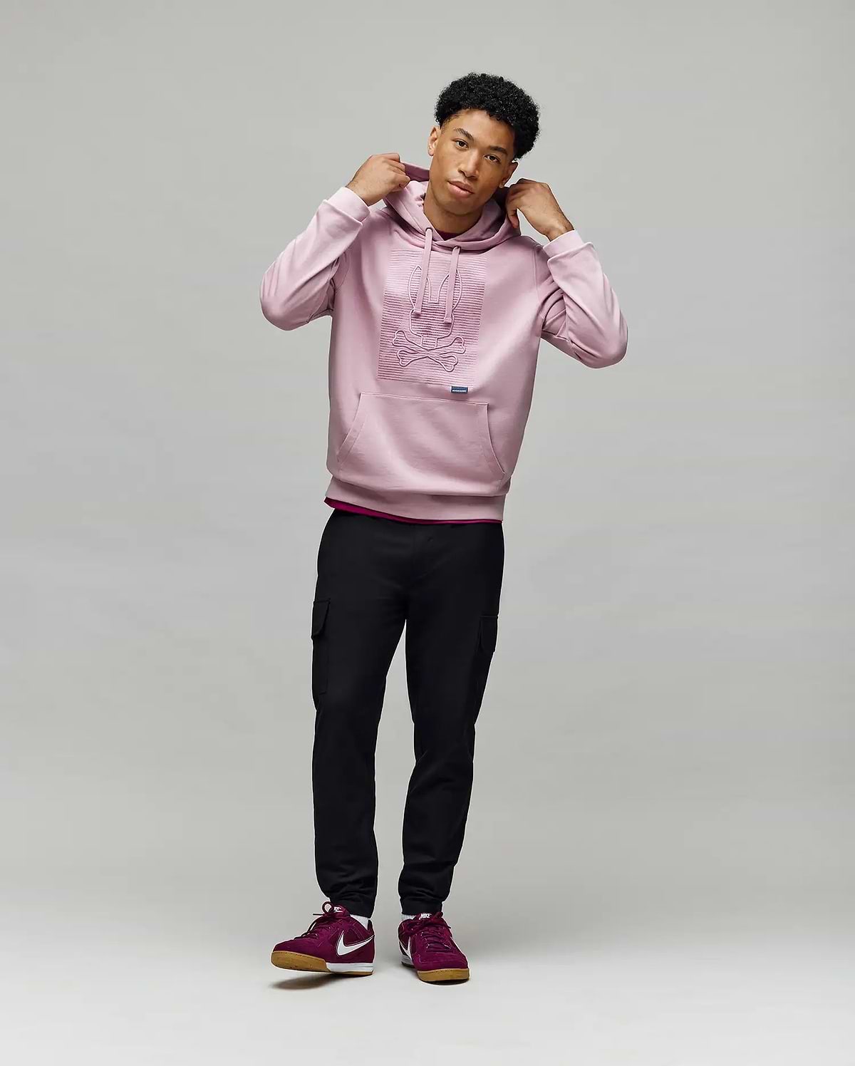 A young man stands against a light background, wearing the Psycho Bunny MENS GRANT HOODIE - MT0500032, black cargo pants, and burgundy Nike sneakers. He holds the hoodie’s drawstrings and gazes slightly to the side.