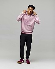 A young man stands against a light background, wearing the Psycho Bunny MENS GRANT HOODIE - MT0500032, black cargo pants, and burgundy Nike sneakers. He holds the hoodie’s drawstrings and gazes slightly to the side.