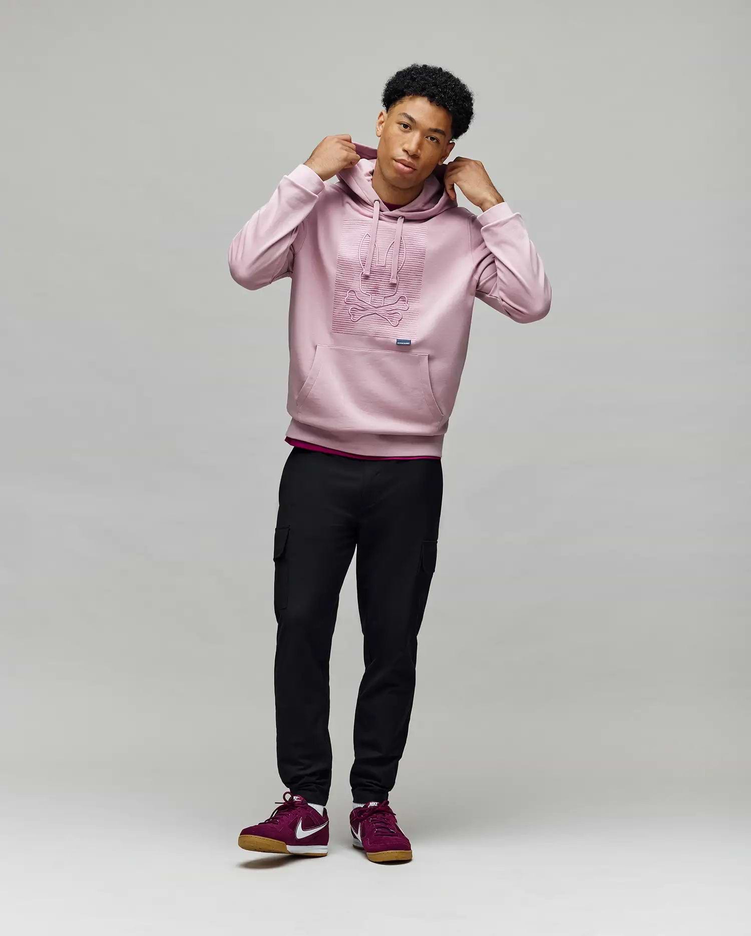 A young man stands against a light background, wearing the Psycho Bunny MENS GRANT HOODIE - MT0500032, black cargo pants, and burgundy Nike sneakers. He holds the hoodie’s drawstrings and gazes slightly to the side.