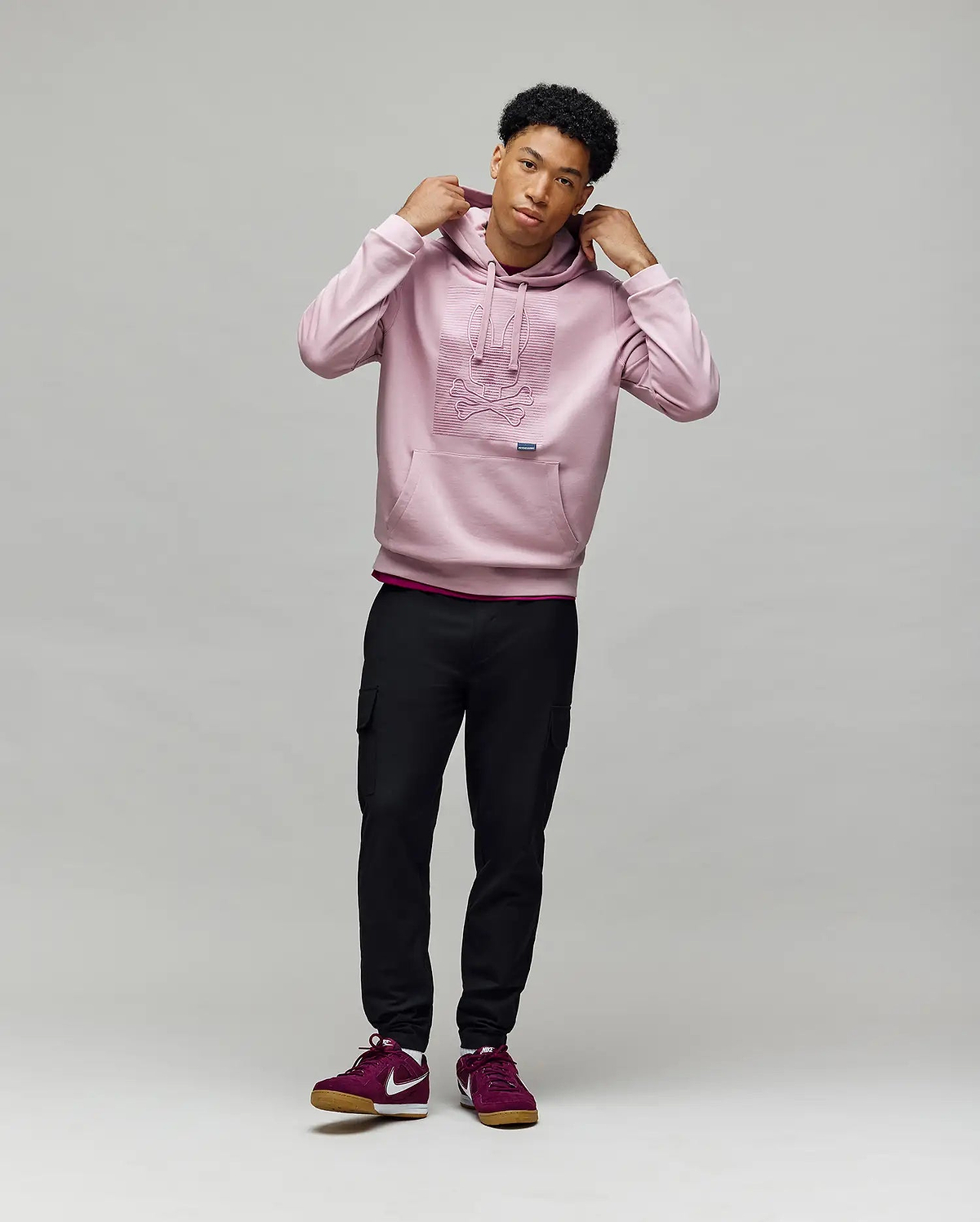 A young man stands against a light background, wearing the Psycho Bunny MENS GRANT HOODIE - MT0500032, black cargo pants, and burgundy Nike sneakers. He holds the hoodie’s drawstrings and gazes slightly to the side.