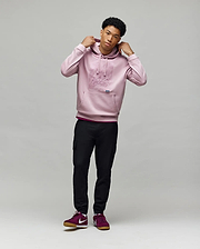 A young man stands against a light background, wearing the Psycho Bunny MENS GRANT HOODIE - MT0500032, black cargo pants, and burgundy Nike sneakers. He holds the hoodie’s drawstrings and gazes slightly to the side.