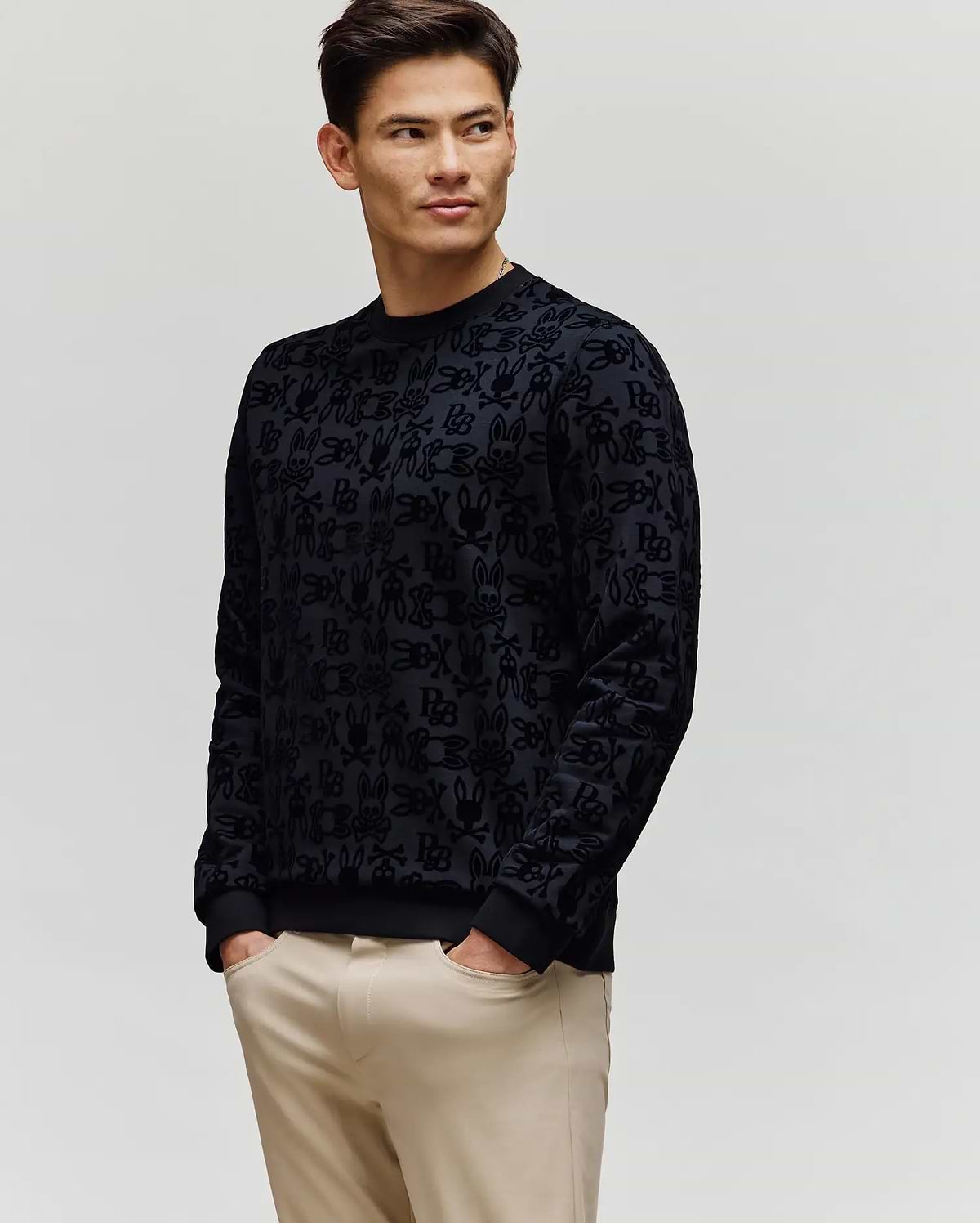 A man wearing the Psycho Bunny MENS REED FLOCK CREW (MT0500041), a black patterned premium cotton blend long-sleeve shirt, with light beige pants stands against a plain light background, looking left with hands in his pockets.