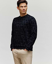 A man wearing the Psycho Bunny MENS REED FLOCK CREW (MT0500041), a black patterned premium cotton blend long-sleeve shirt, with light beige pants stands against a plain light background, looking left with hands in his pockets.