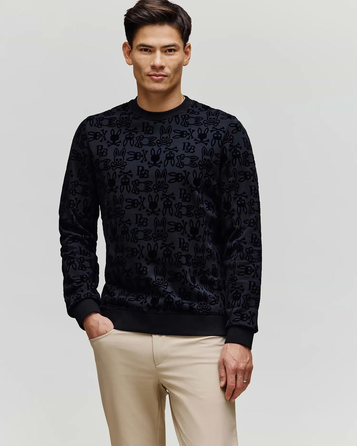 A man with short dark hair wears Psycho Bunny's MENS REED FLOCK CREW (MT0500041), a black premium cotton blend pullover sweater, and beige pants, standing with one hand in his pocket against a plain light background.