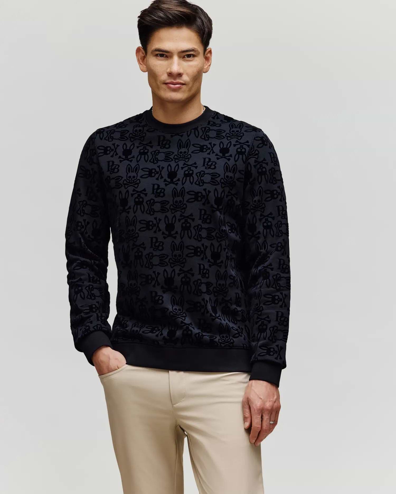 A man with short dark hair wears Psycho Bunny's MENS REED FLOCK CREW (MT0500041), a black premium cotton blend pullover sweater, and beige pants, standing with one hand in his pocket against a plain light background.