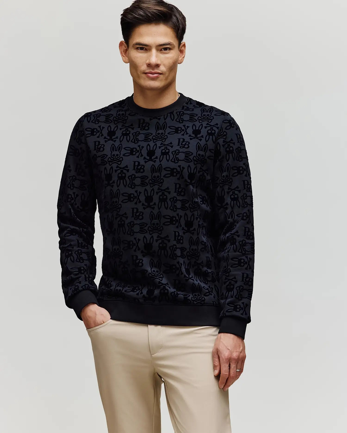 A man with short dark hair wears Psycho Bunny's MENS REED FLOCK CREW (MT0500041), a black premium cotton blend pullover sweater, and beige pants, standing with one hand in his pocket against a plain light background.