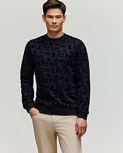A man with short dark hair wears Psycho Bunny's MENS REED FLOCK CREW (MT0500041), a black premium cotton blend pullover sweater, and beige pants, standing with one hand in his pocket against a plain light background.