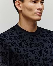 A man models the Psycho Bunny MENS REED FLOCK CREW (MT0500041), featuring skulls, crossbones, and bunny ears on a crewneck pullover. Shown from shoulders up against a neutral background. Made from a premium cotton blend for comfort.