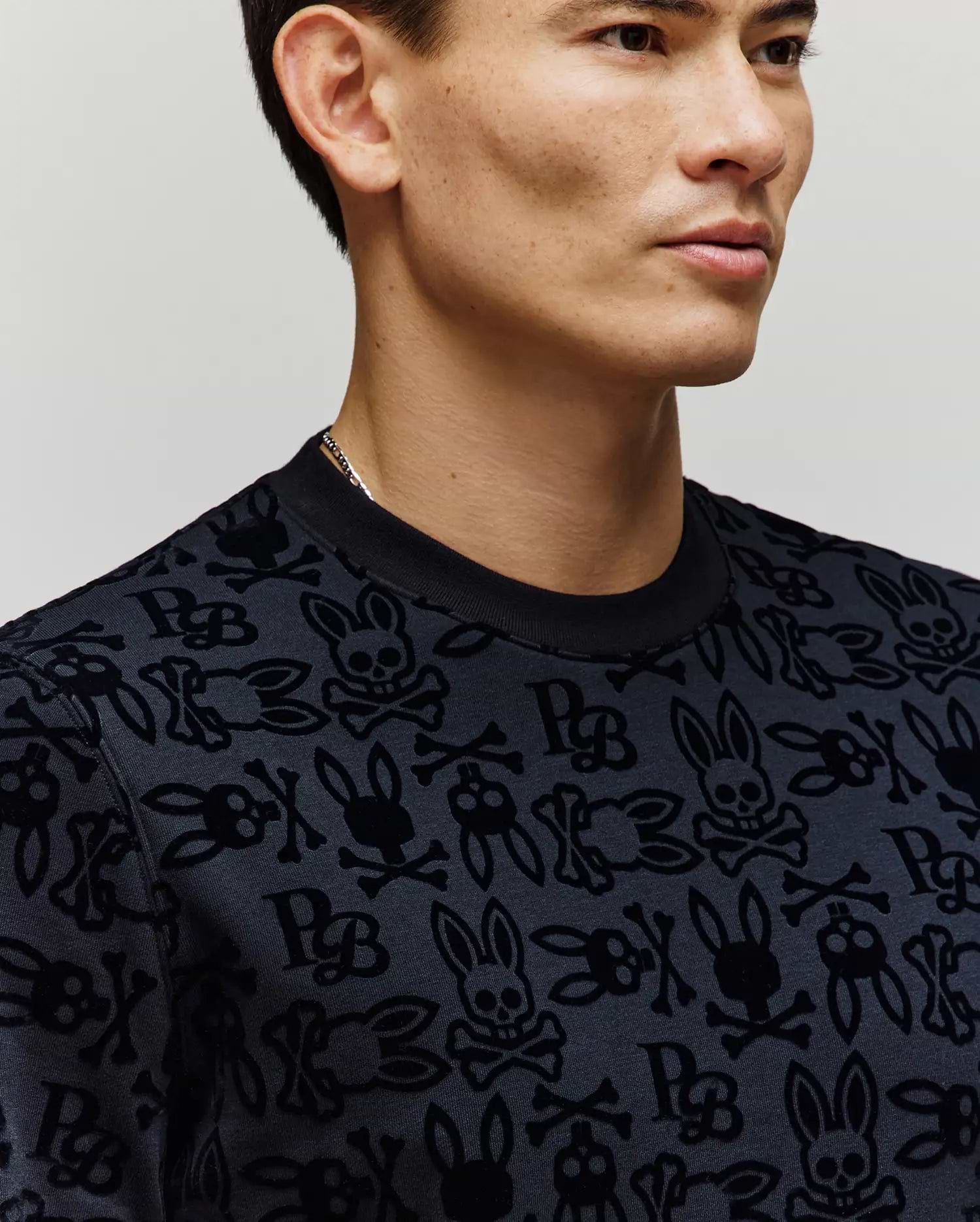 A man models the Psycho Bunny MENS REED FLOCK CREW (MT0500041), featuring skulls, crossbones, and bunny ears on a crewneck pullover. Shown from shoulders up against a neutral background. Made from a premium cotton blend for comfort.