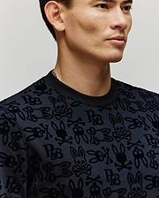 A man models the Psycho Bunny MENS REED FLOCK CREW (MT0500041), featuring skulls, crossbones, and bunny ears on a crewneck pullover. Shown from shoulders up against a neutral background. Made from a premium cotton blend for comfort.