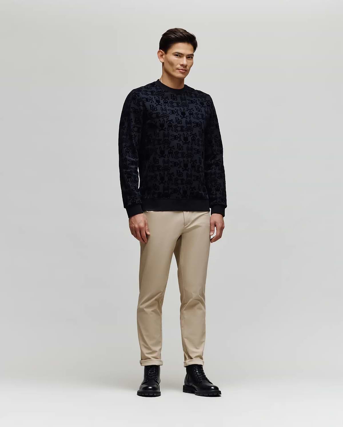 A man stands against a plain light gray background, wearing the Psycho Bunny MENS REED FLOCK CREW (MT0500041) pullover sweater, paired with beige pants and black boots, facing slightly sideways with a neutral expression.