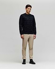 A man stands against a plain light gray background, wearing the Psycho Bunny MENS REED FLOCK CREW (MT0500041) pullover sweater, paired with beige pants and black boots, facing slightly sideways with a neutral expression.