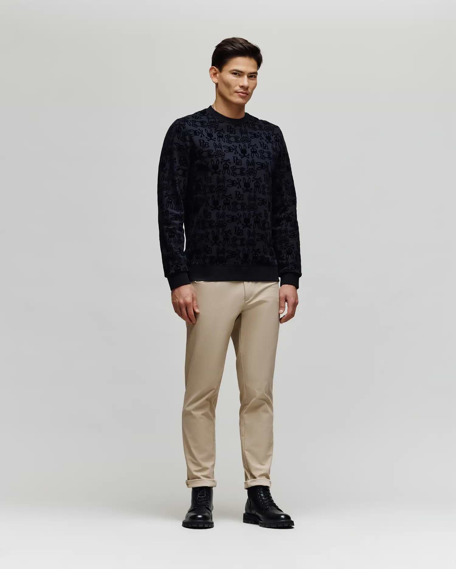A man stands against a plain light gray background, wearing the Psycho Bunny MENS REED FLOCK CREW (MT0500041) pullover sweater, paired with beige pants and black boots, facing slightly sideways with a neutral expression.