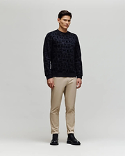 A man stands against a plain light gray background, wearing the Psycho Bunny MENS REED FLOCK CREW (MT0500041) pullover sweater, paired with beige pants and black boots, facing slightly sideways with a neutral expression.