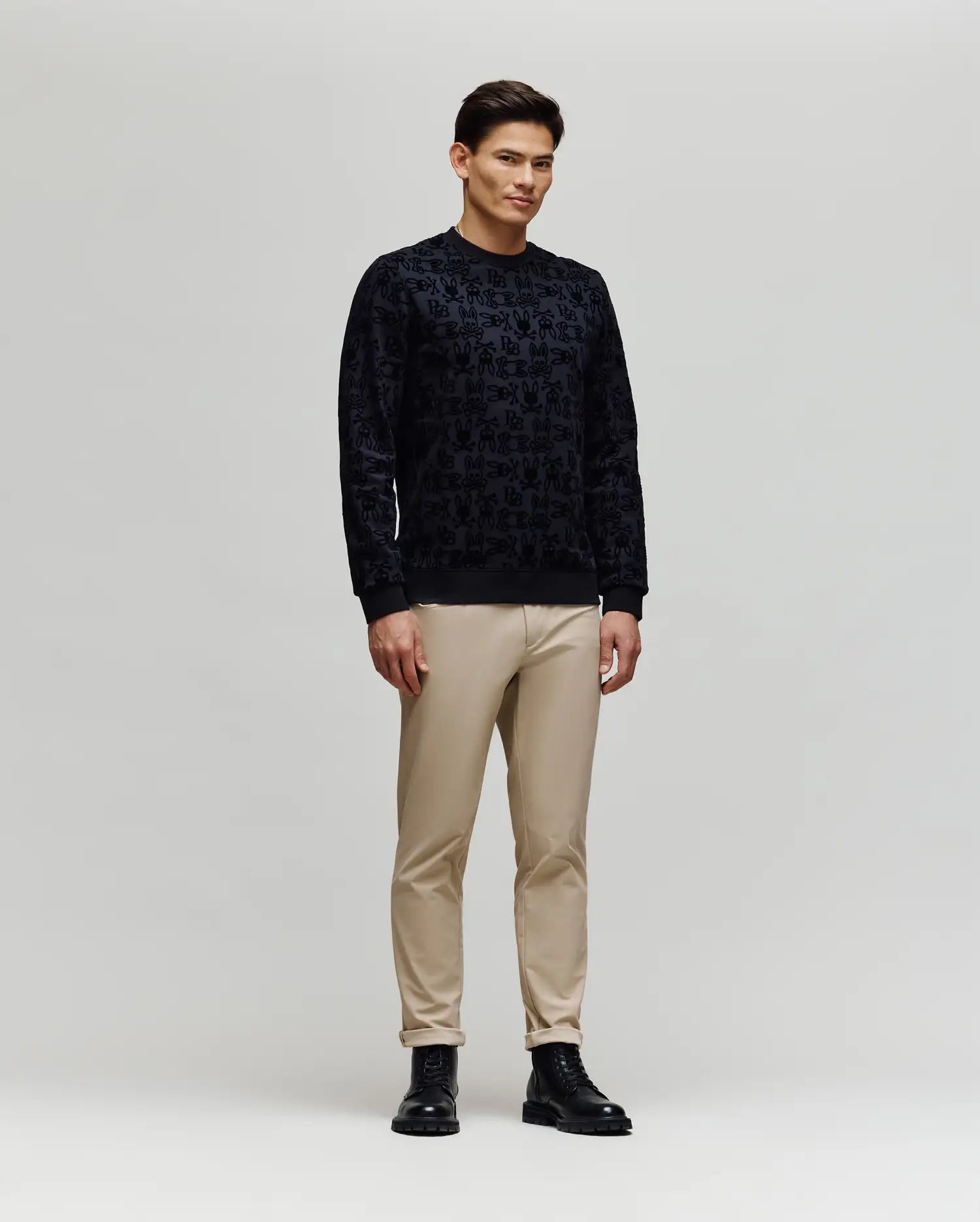 A man stands against a plain light gray background, wearing the Psycho Bunny MENS REED FLOCK CREW (MT0500041) pullover sweater, paired with beige pants and black boots, facing slightly sideways with a neutral expression.