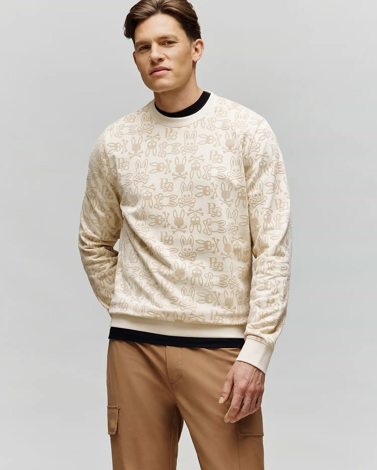 A man with short brown hair models the Psycho Bunny MENS REED FLOCK CREW - MT0500041, a premium cotton blend pullover with a beige hand sign pattern, layered over a black shirt and tan cargo pants, against a plain light gray background.