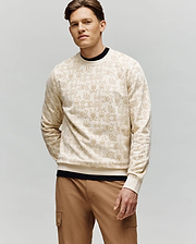 A man with short brown hair models the Psycho Bunny MENS REED FLOCK CREW - MT0500041, a premium cotton blend pullover with a beige hand sign pattern, layered over a black shirt and tan cargo pants, against a plain light gray background.