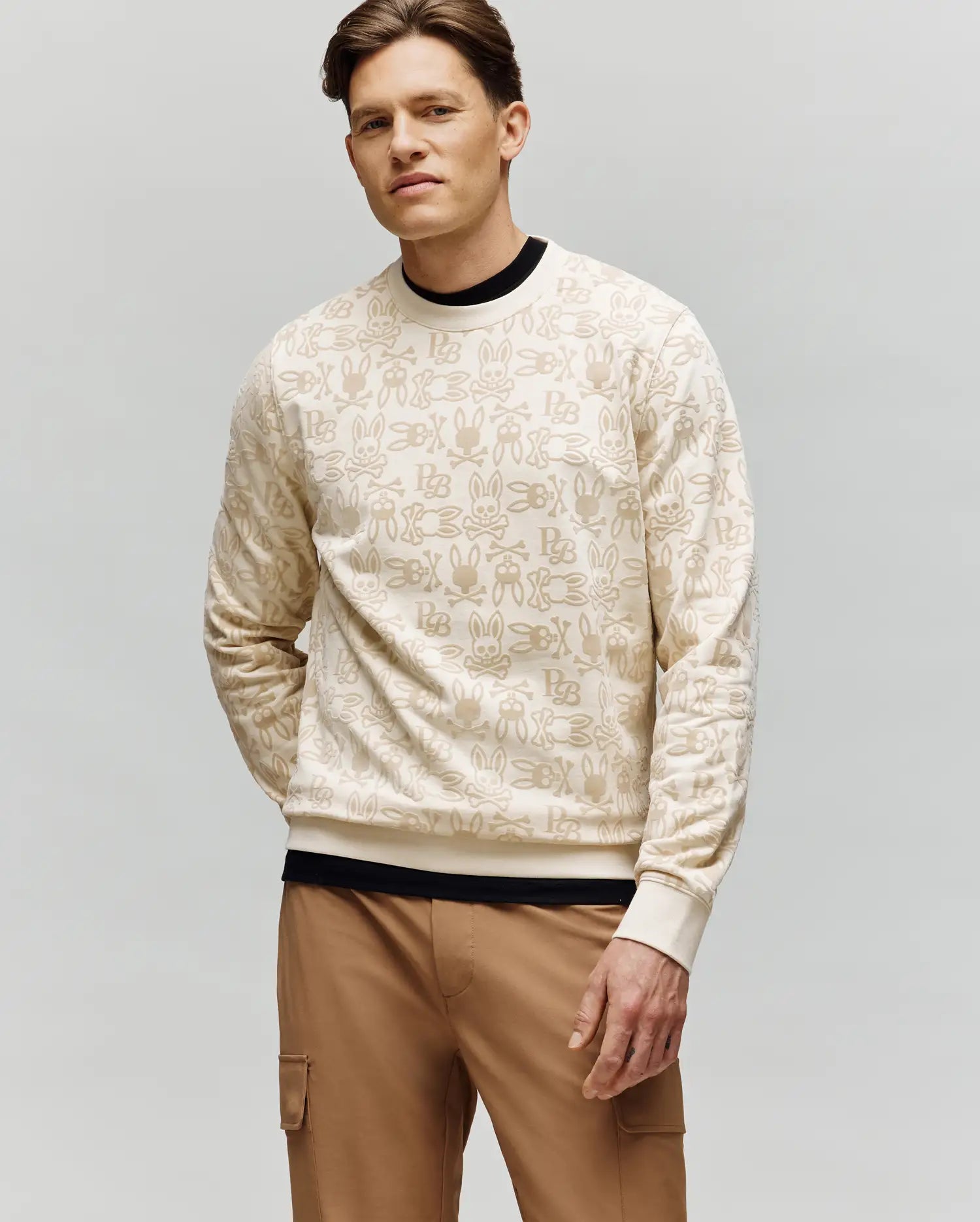 A man with short brown hair models the Psycho Bunny MENS REED FLOCK CREW - MT0500041, a premium cotton blend pullover with a beige hand sign pattern, layered over a black shirt and tan cargo pants, against a plain light gray background.