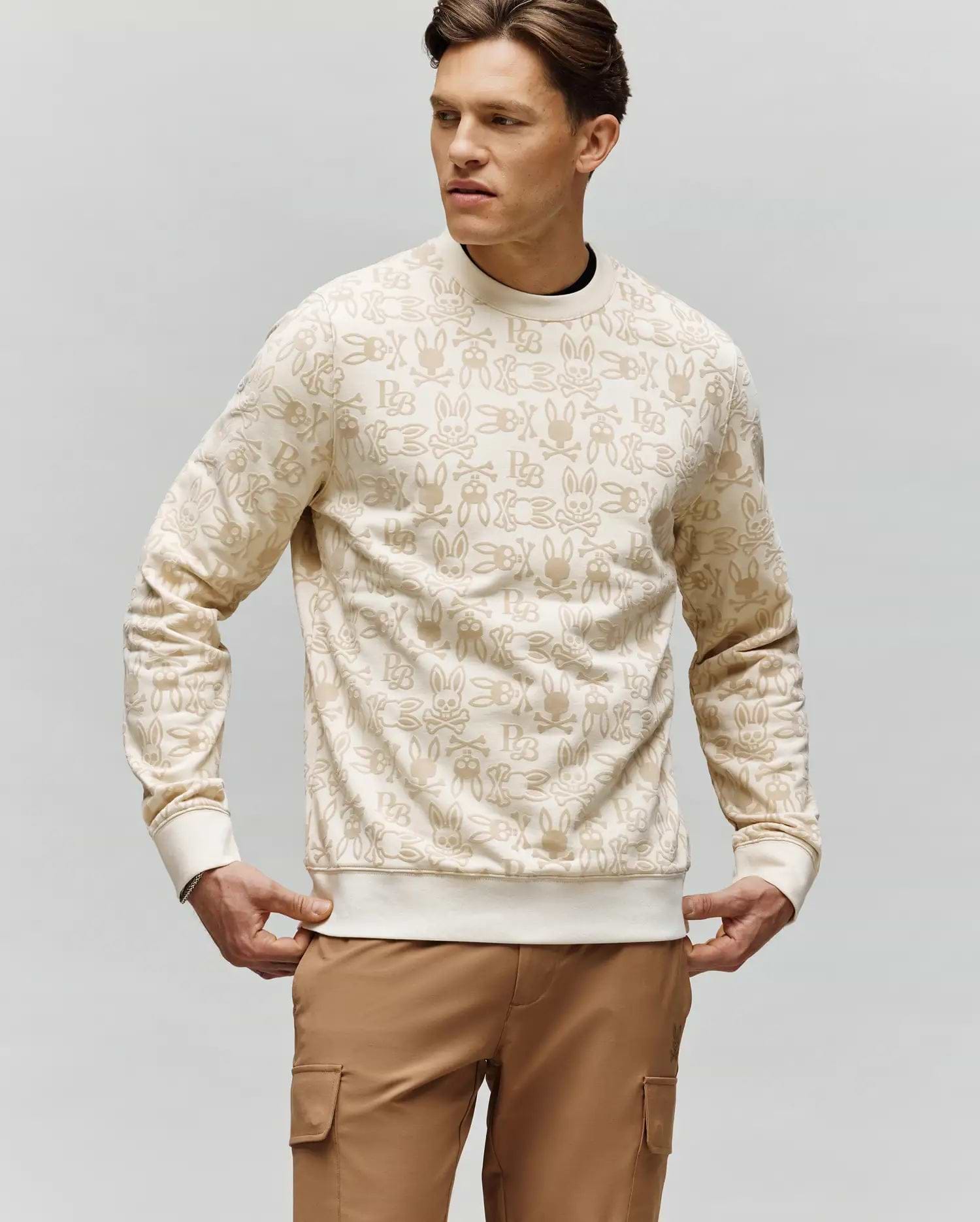 A man wears the Psycho Bunny MENS REED FLOCK CREW - MT0500041 with bunny print and tan cargo pants, standing with hands by his sides and looking left against a plain background.