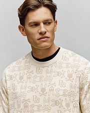 A man with short brown hair models the Psycho Bunny MENS REED FLOCK CREW - MT0500041, a cream crewneck pullover with an all-over cartoon bunny and PB pattern. He gazes to the side against a plain, light gray background.