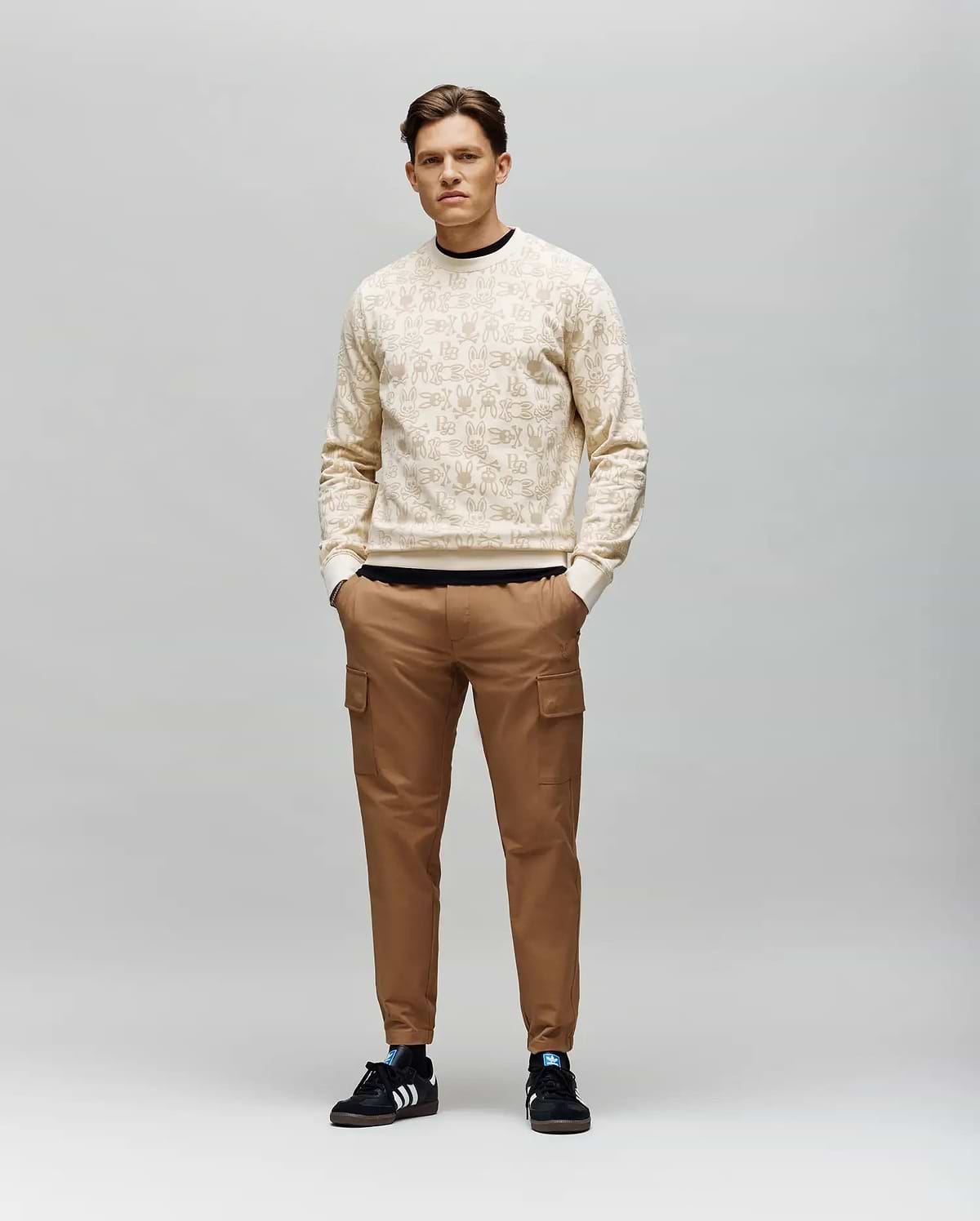 A man stands against a gray background wearing the Psycho Bunny MENS REED FLOCK CREW - MT0500041, a light beige premium cotton blend pullover sweater. He pairs it with brown cargo pants and black Adidas sneakers with white stripes, hands in his pockets.