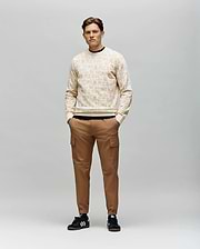A man stands against a gray background wearing the Psycho Bunny MENS REED FLOCK CREW - MT0500041, a light beige premium cotton blend pullover sweater. He pairs it with brown cargo pants and black Adidas sneakers with white stripes, hands in his pockets.