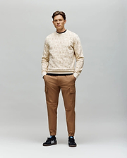 A man stands against a gray background wearing the Psycho Bunny MENS REED FLOCK CREW - MT0500041, a light beige premium cotton blend pullover sweater. He pairs it with brown cargo pants and black Adidas sneakers with white stripes, hands in his pockets.