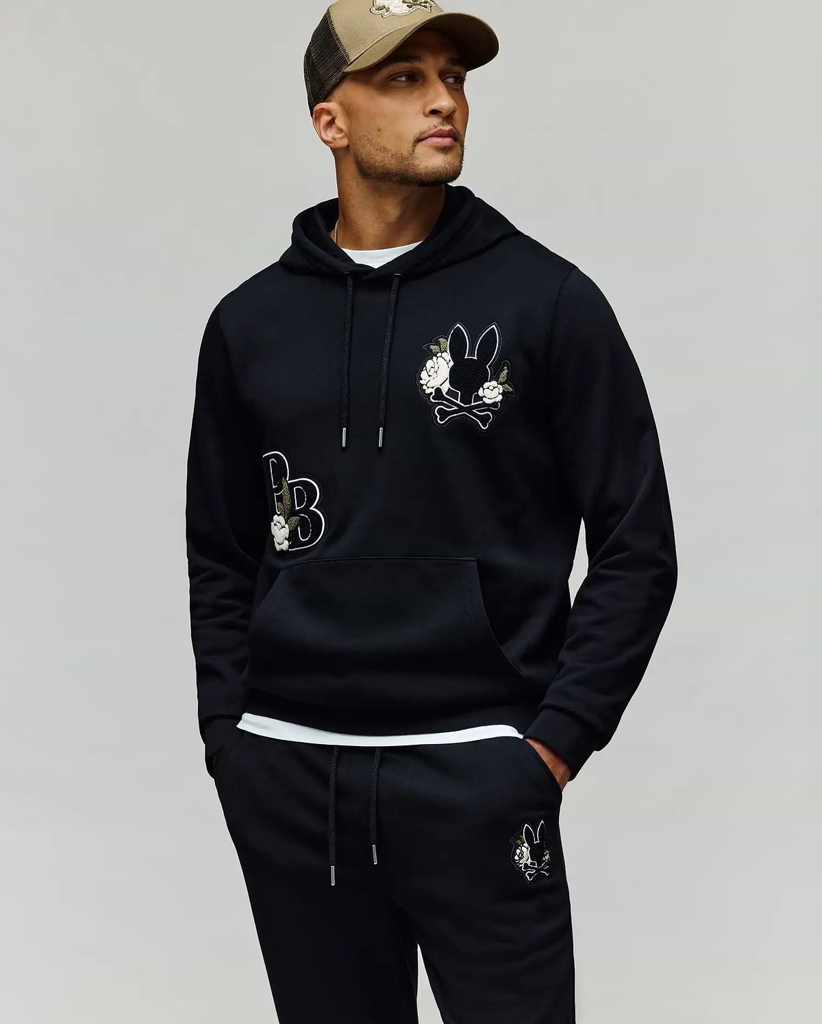 A man wears the Psycho Bunny MENS AUGUSTE HOODIE - MT0500043 in black with cartoon bunny patches and chenille floral details, matching sweatpants, and a beige cap, standing hands-in-pockets against a plain background.